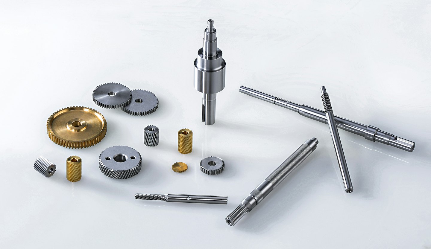 Ingranaggi e alberi metallici assortiti disposti su una superficie bianca.